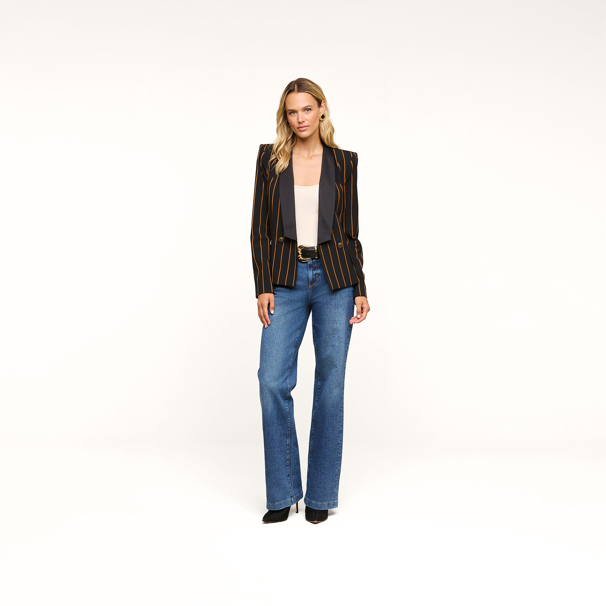 A woman stands facing forward against a plain white background, wearing the Imogen Satin Lapel Blazer—a black double-breasted blazer with a satin lapel—paired with a light gray top, blue jeans, and black shoes.