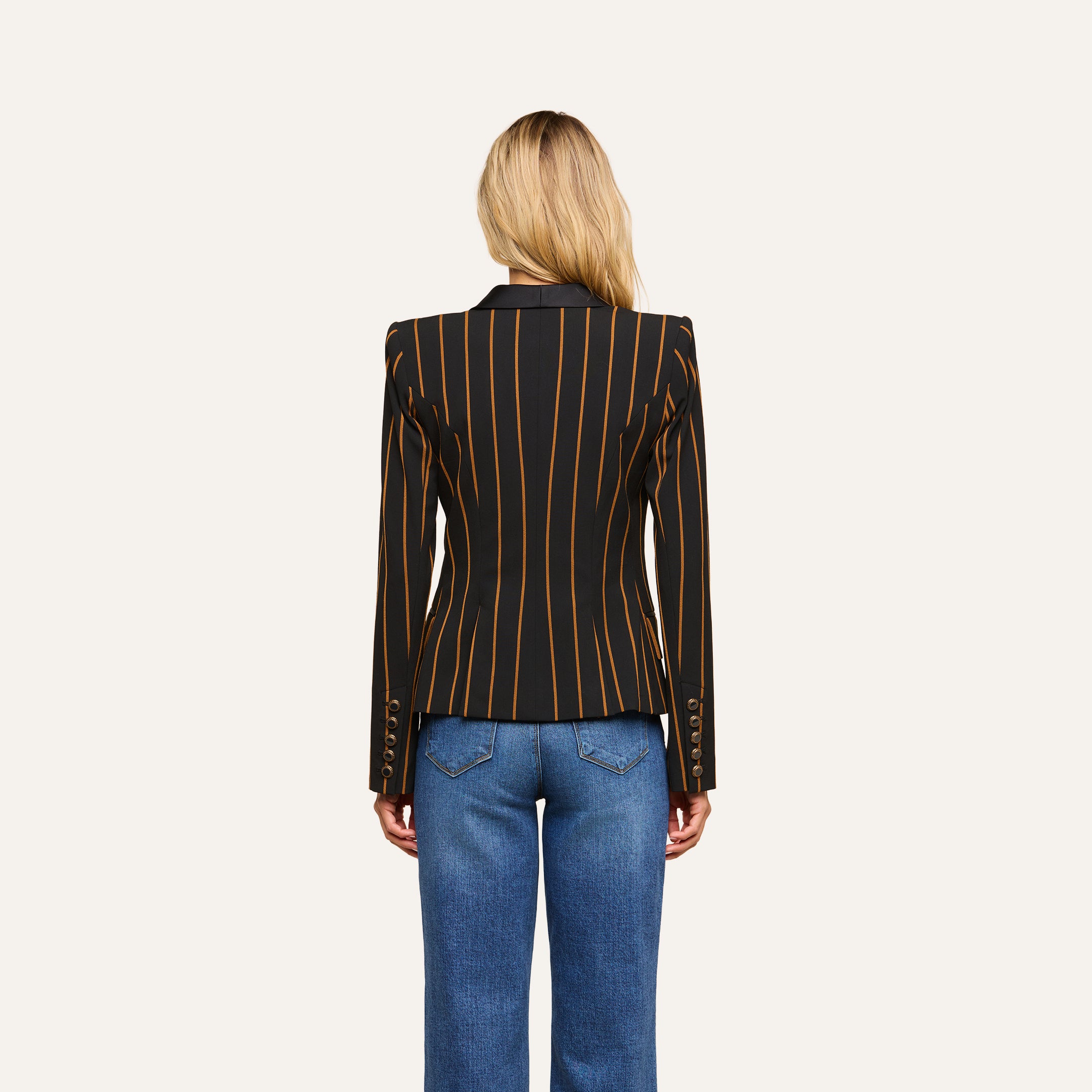 A person with long blond hair, seen from behind, wears the Imogen Satin Lapel Blazer—a black double-breasted blazer with gold vertical stripes—paired with blue jeans, standing against a plain white background.