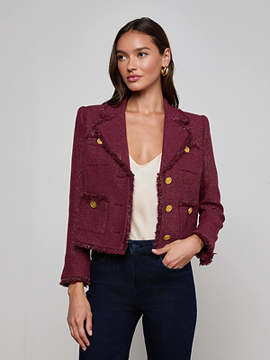 A woman with straight brown hair looks slightly to the side, wearing the L'AGENCE Sylvia Tweed Jacket with gold buttons and fringed trims over a cream top and dark jeans, set against a plain light gray background.