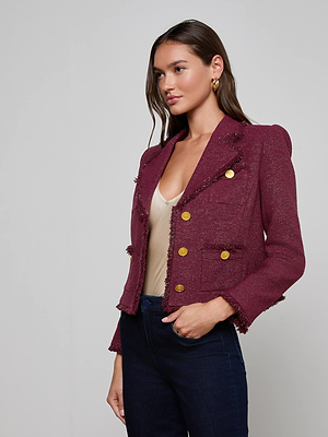 A woman stands against a plain light background, looking slightly to the side, wearing the Sylvia Tweed Jacket by L'AGENCE with gold buttons and fringed trims, paired with a beige top and dark jeans.