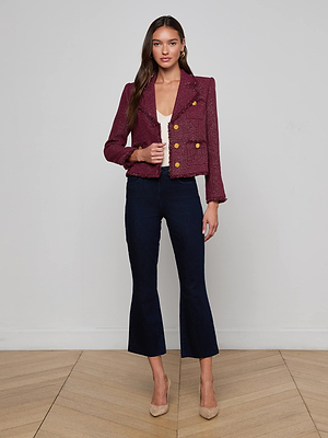 A woman with long brown hair and a neutral expression stands against a plain wall, wearing the L'AGENCE Sylvia Tweed Jacket with gold buttons, a white top, dark blue cropped pants, and beige heels on a wood-patterned floor.