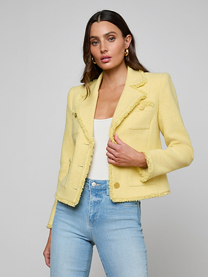 A woman stands against a plain gray background, wearing the Sylvia Tweed Jacket in light yellow with fringed trims and gold buttons, layered over a white top and paired with light blue high-waisted jeans.