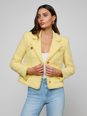 A woman wears the Sylvia Tweed Jacket—a pale yellow tweed piece with gold buttons and frayed edges—layered over a white top and light blue jeans, standing against a plain light gray background.