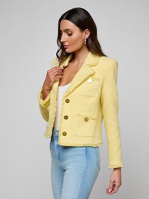 A woman with long brown hair wears the Sylvia Tweed Jacket in light yellow, featuring gold buttons, over a white top and light blue jeans. She stands sideways, looking down as her hand touches the jacket’s collar.