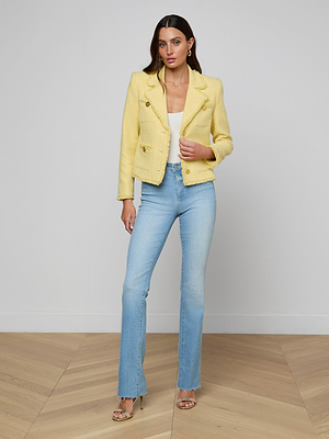A woman with long dark hair poses indoors against a light background and wooden floor, wearing the Sylvia Tweed Jacket—yellow, fitted with gold buttons—paired with a white top, light blue jeans, heeled sandals, and minimal accessories.