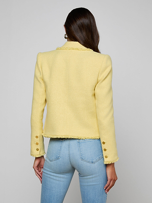 A woman with long brown hair, facing away, wears the Sylvia Tweed Jacket in pale yellow with gold sleeve buttons and light blue jeans, set against a plain gray background.