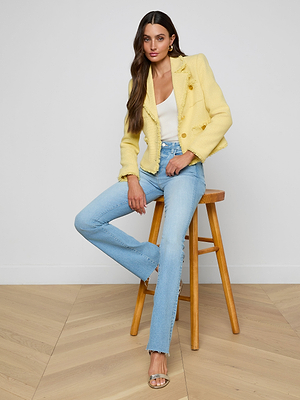 A woman with long brown hair sits on a wooden stool, wearing the Sylvia Tweed Jacket in yellow with gold buttons, paired with a white top, light blue jeans, and transparent heeled sandals against a plain white backdrop with a light wood floor.