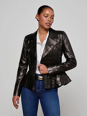 A woman wears the L'AGENCE Lyric Leather Peplum Jacket in shiny black lambskin over a white collared shirt and blue jeans, standing against a light background and looking to the side with one hand on her jacket.