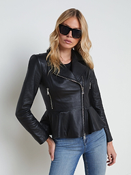 A woman wears the Lyric Leather Peplum Jacket—crafted in black lambskin leather with zip details—paired with blue jeans and dark sunglasses, as she stands against a plain light background.