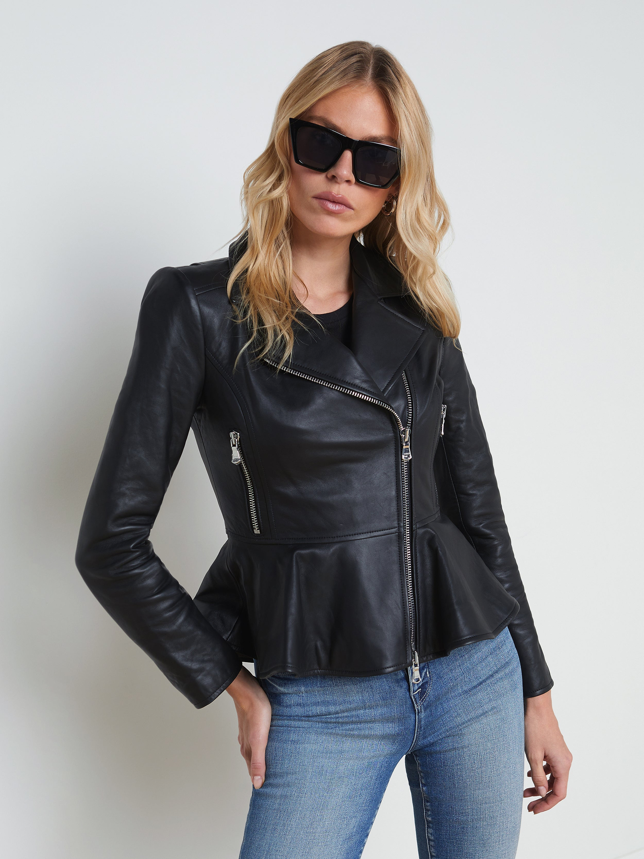 A woman wears the Lyric Leather Peplum Jacket—crafted in black lambskin leather with zip details—paired with blue jeans and dark sunglasses, as she stands against a plain light background.