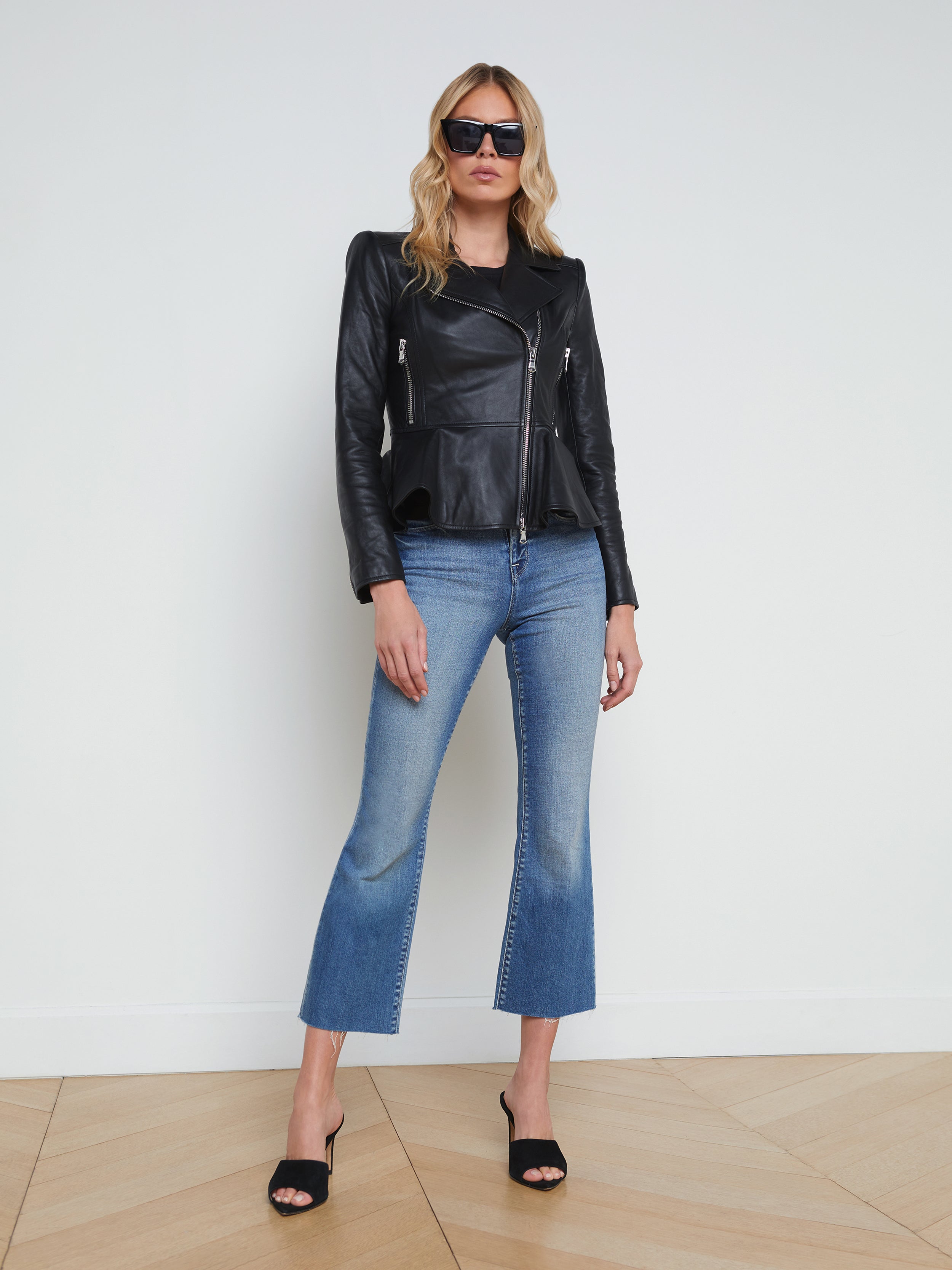 A woman wearing sunglasses, the Lyric Leather Peplum Jacket in black lambskin, blue cropped jeans, and black heels stands against a plain white background.
