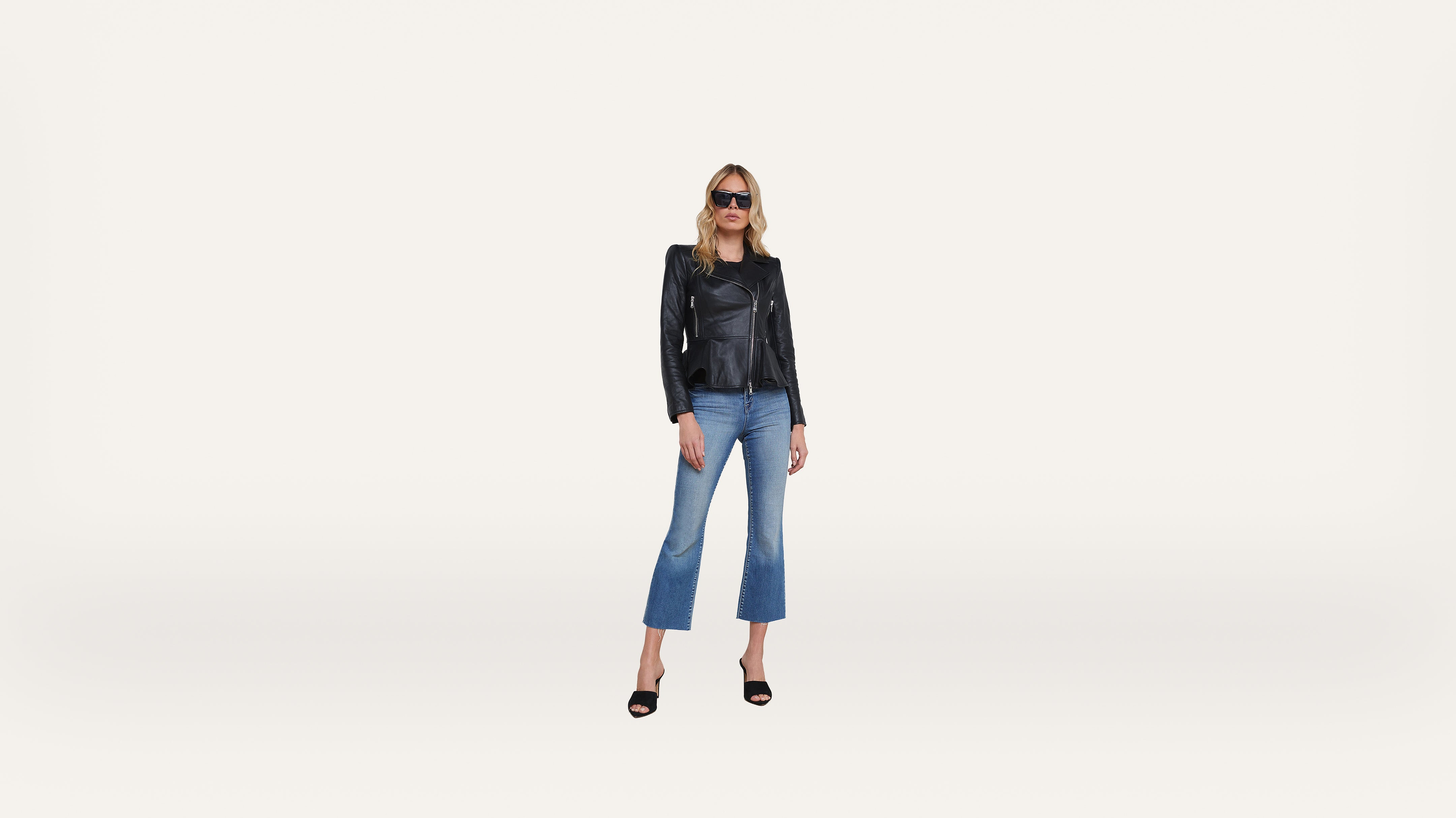 A woman wearing sunglasses, a black leather jacket, blue cropped jeans, and black heels stands confidently against a plain white background.