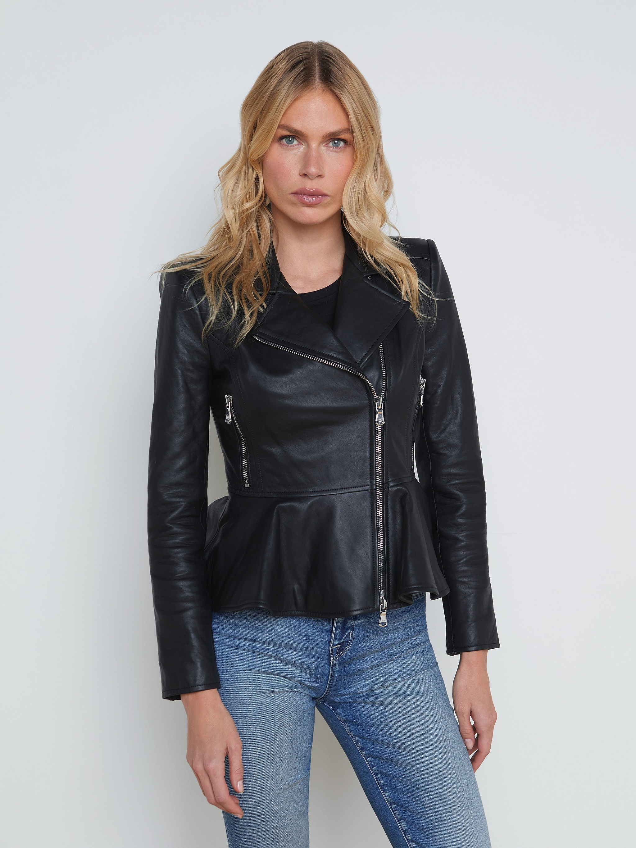 A woman with wavy blonde hair wears the Lyric Leather Peplum Jacket in black lambskin leather and blue jeans, standing against a plain off-white background, facing forward with a neutral expression.