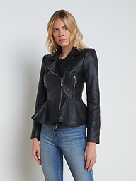 A woman wearing the Lyric Leather Peplum Jacket—crafted from black lambskin leather with a peplum hem and silver zippers—is paired with blue jeans, standing against a plain light background.