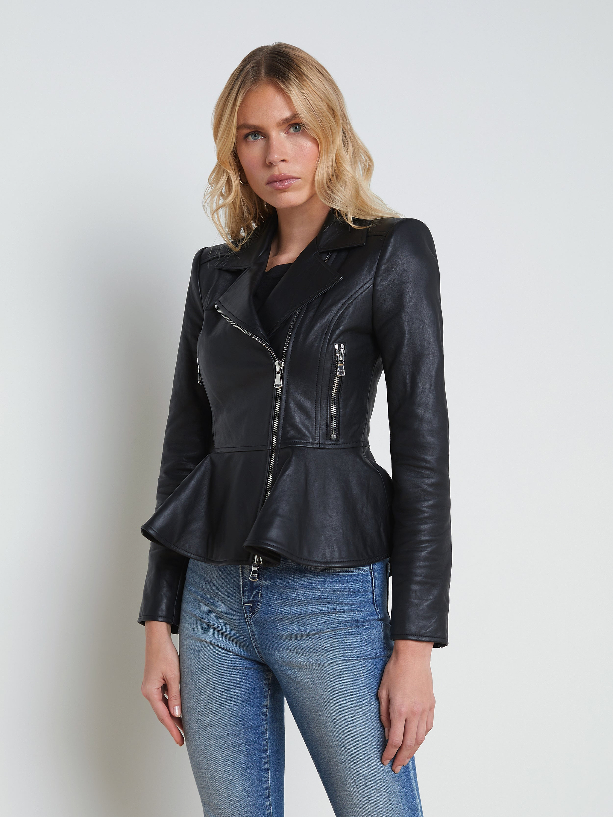 A woman wearing the Lyric Leather Peplum Jacket—crafted from black lambskin leather with a peplum hem and silver zippers—is paired with blue jeans, standing against a plain light background.