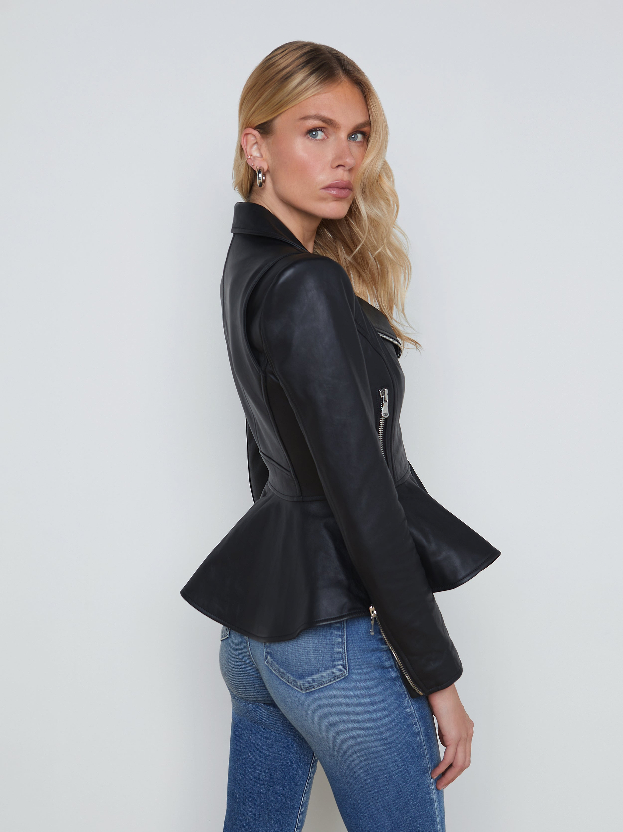 A woman with long blonde hair stands sideways against a plain light background, wearing the Lyric Leather Peplum Jacket in black lambskin over blue jeans.