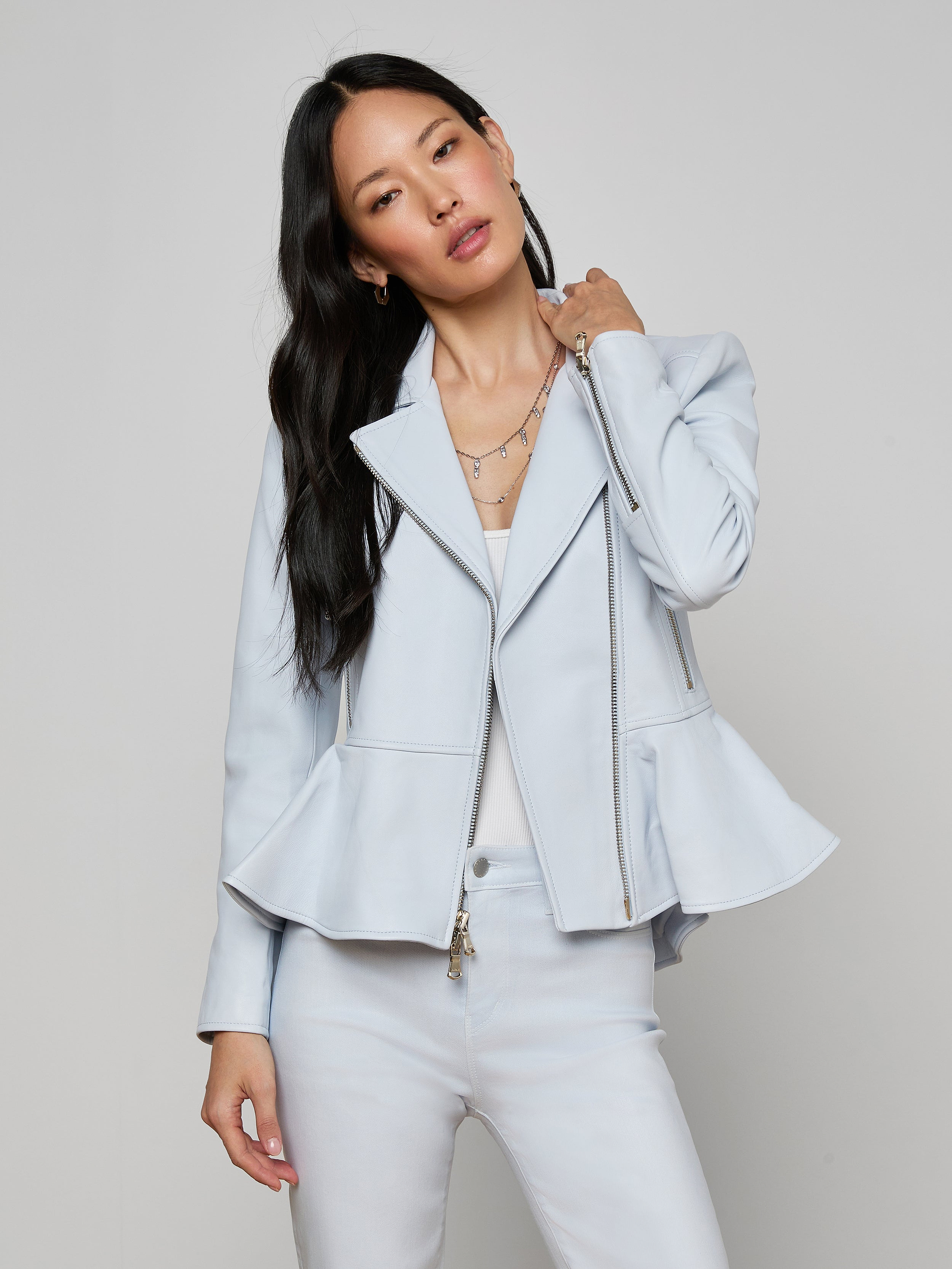 A woman stands against a plain background, looking at the camera and holding her hair with one hand while wearing the Lyric Peplum Biker Jacket in powder blue over a white top and matching pants.
