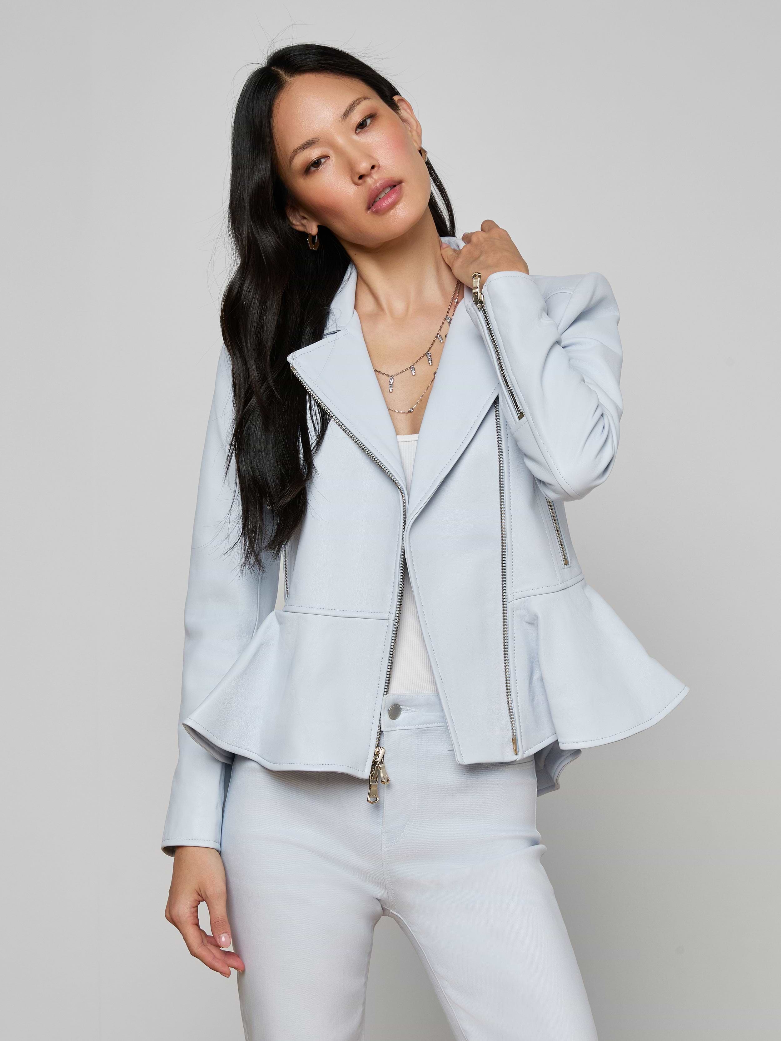 A woman stands against a plain background, looking at the camera and holding her hair with one hand while wearing the Lyric Peplum Biker Jacket in powder blue over a white top and matching pants.