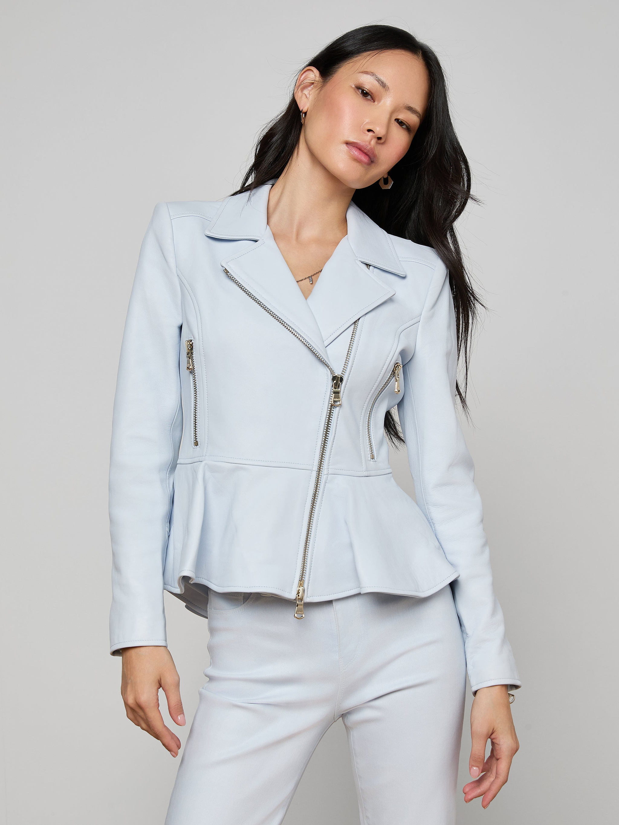 A woman with long dark hair wears the Lyric Peplum Biker Jacket in powder blue, featuring zipper details, along with matching pants, standing against a plain light background.