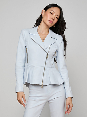 A woman with long dark hair wears the Lyric Peplum Biker Jacket in light blue, featuring zipper details and a subtle peplum hem, paired with matching pants and posing confidently against a plain light gray background.