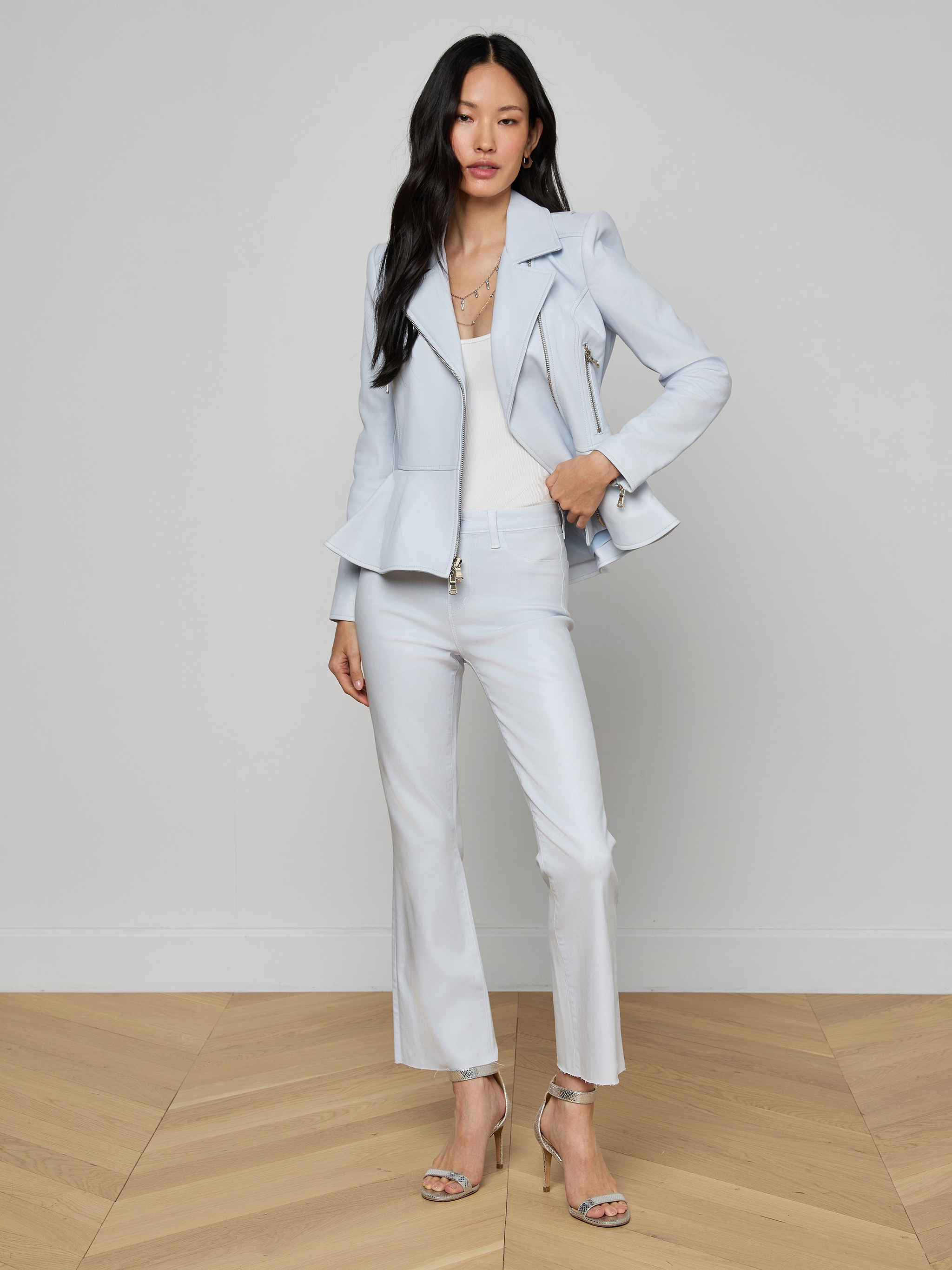 A woman with long black hair wears the Lyric Peplum Biker Jacket in powder blue and cropped pants, paired with strappy high-heeled sandals, standing against a plain white background.