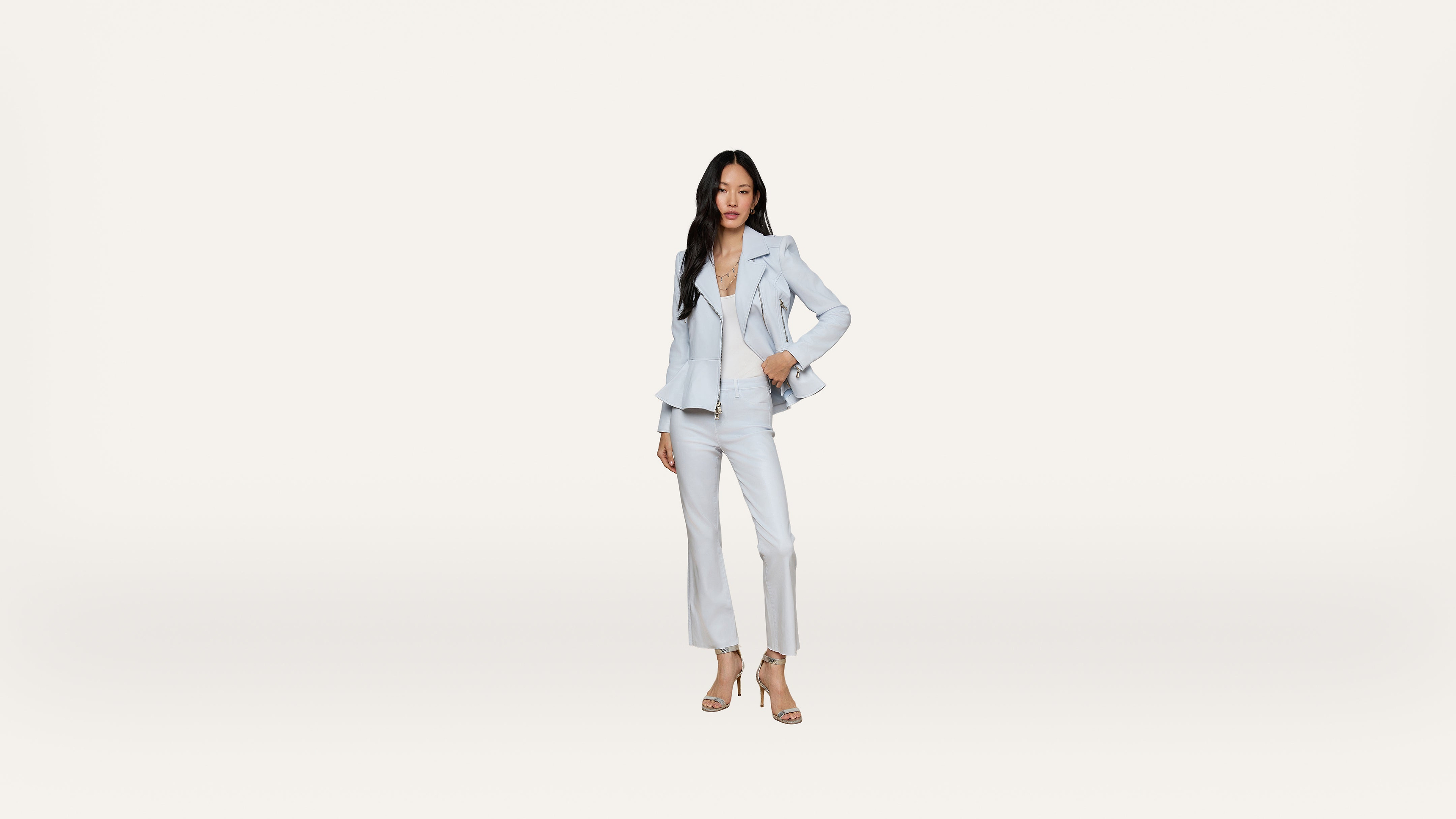 Woman in a light blue suit and heels stands confidently against a plain white background, one hand on her hip and the other relaxed by her side.