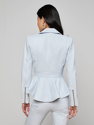A woman with long dark hair is seen from behind, wearing the Lyric Peplum Biker Jacket in light blue with a peplum hem and gold zipper details, paired with matching pants for a chic, feminine take on classic biker style.