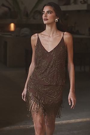A woman wearing a sleeveless, brown dress with gold embroidery and fringe detailing stands indoors. She has long hair, gold earrings, and is looking slightly to the side with a soft expression.