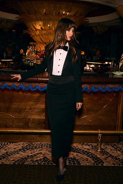 A woman in a black tuxedo-style dress with a bow tie stands by an ornate bar, looking to the side. The bar has intricate woodwork and bottles in the background, creating an elegant, dimly lit atmosphere.