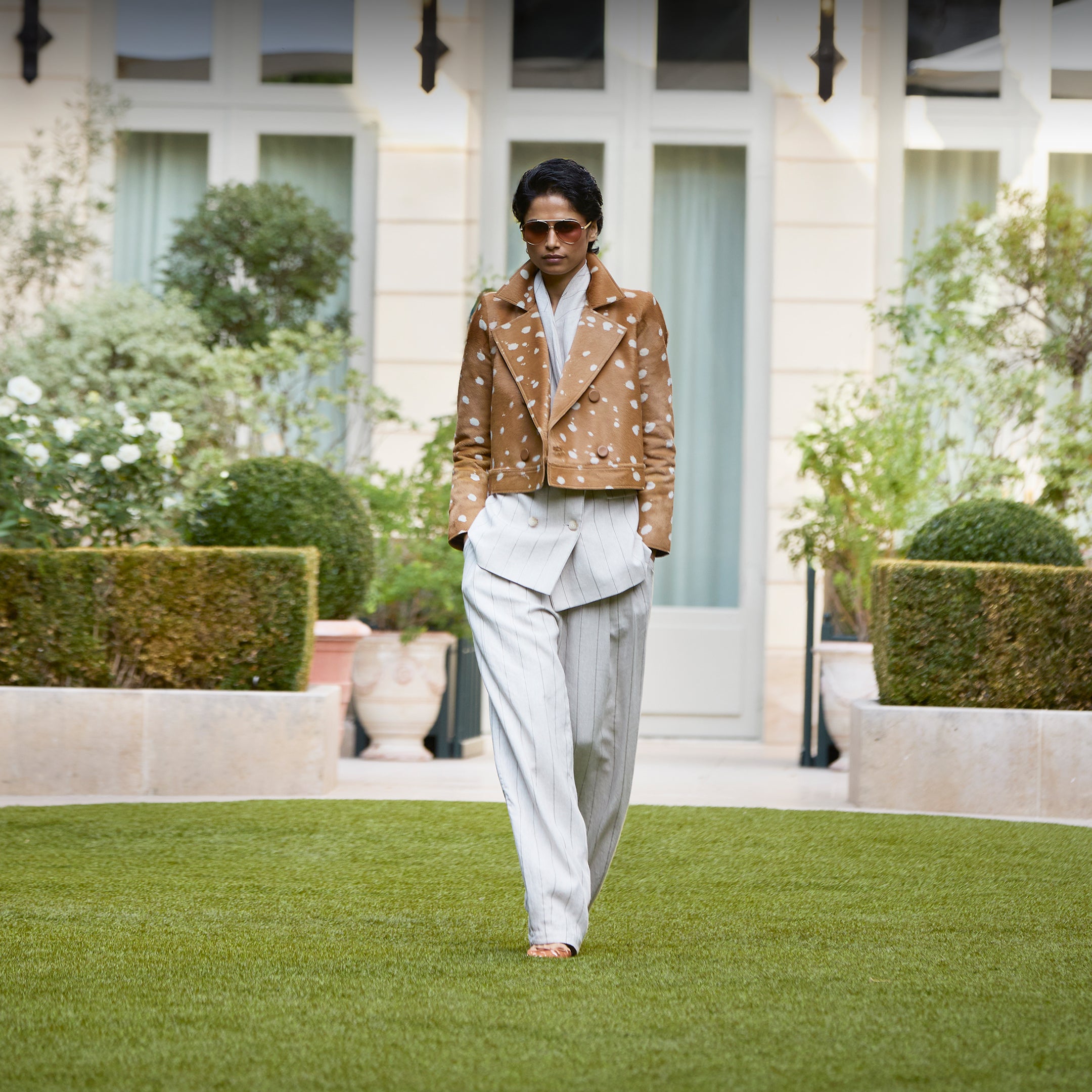 A model walks on a manicured green lawn in front of a grand villa, wearing a brown cropped jacket with a white floral print and light gray pinstriped trousers.