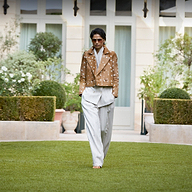 A model walks on a manicured green lawn in front of a grand villa, wearing a brown cropped jacket with a white floral print and light gray pinstriped trousers.
