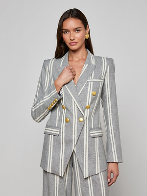 A woman with long brown hair and gold earrings stands against a plain background, wearing the Kendi Longline Blazer—a gray double-breasted style featuring white vertical stripes, gold buttons, and holding its lapel.
