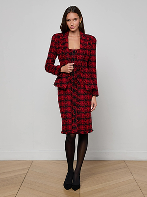 A woman with long brown hair and a neutral expression stands against a plain light wall, wearing the L'AGENCE Kaisley Tweed Blazer in red and black houndstooth, styled with a matching skirt, black tights, and heels.