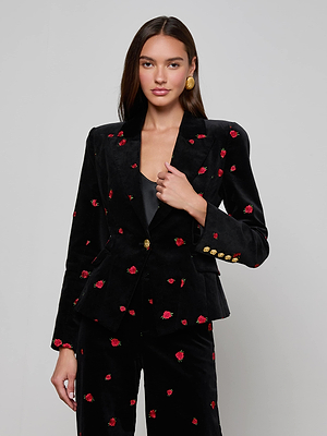 A woman poses against a plain background wearing the Kaisley Velvet Blazer by L'AGENCE—black with red floral embroidery, gold buttons, statement earrings, and a black top underneath.