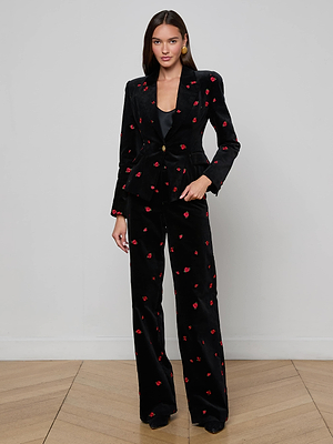A woman with straight brown hair poses against a white wall on a wooden floor, wearing the Kaisley Velvet Blazer by L'AGENCE featuring black velvet with red floral embroidery and wide-leg pants.