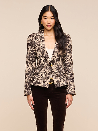 A woman with long, dark hair stands against a plain beige background, facing forward with a neutral expression. She is wearing the Kaisley Blazer, which features an antique-inspired print, layered over a white top and paired with dark pants.
