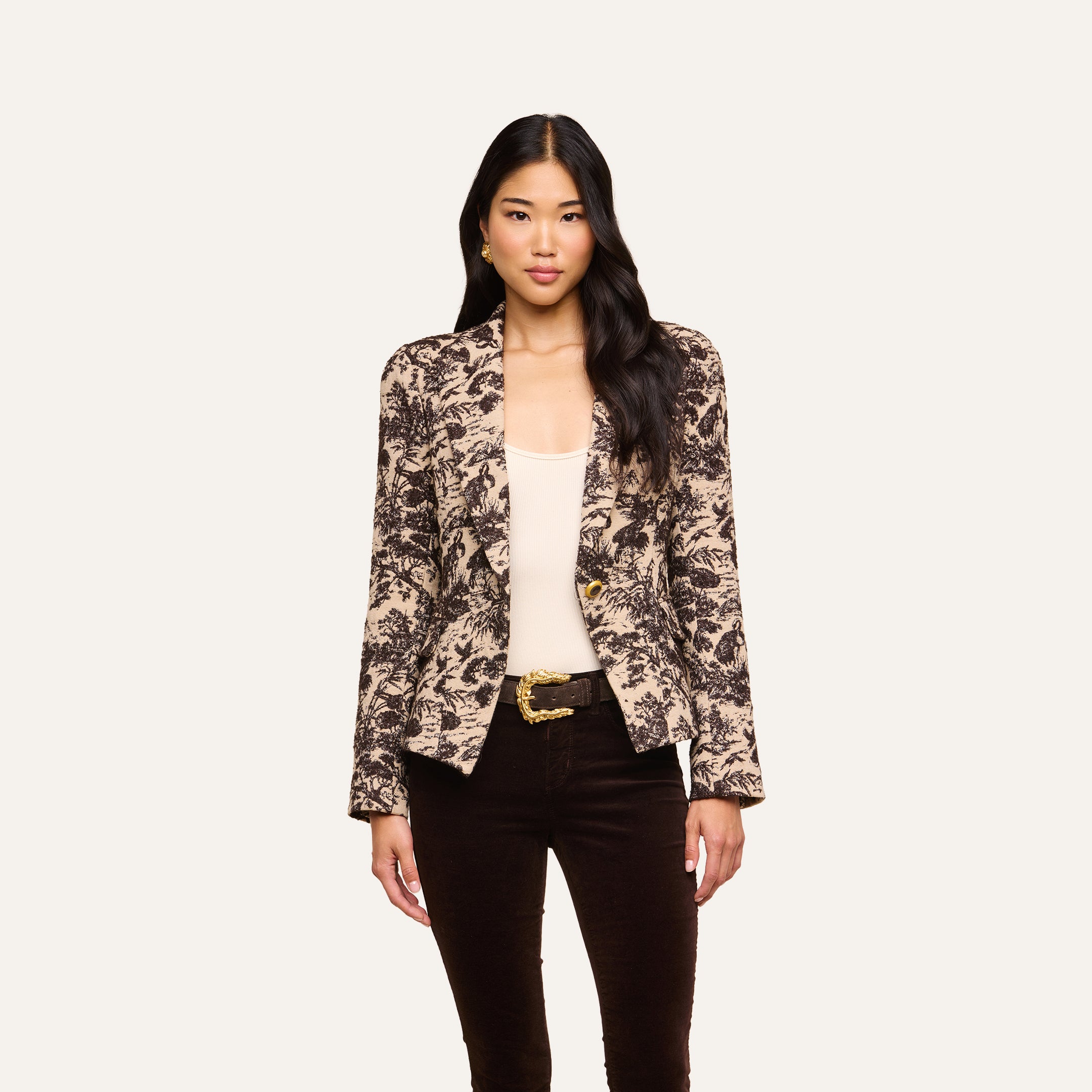 A woman stands facing forward, dressed in the Kaisley Blazer featuring an antique-inspired print, layered over a light top and dark pants against a plain background.