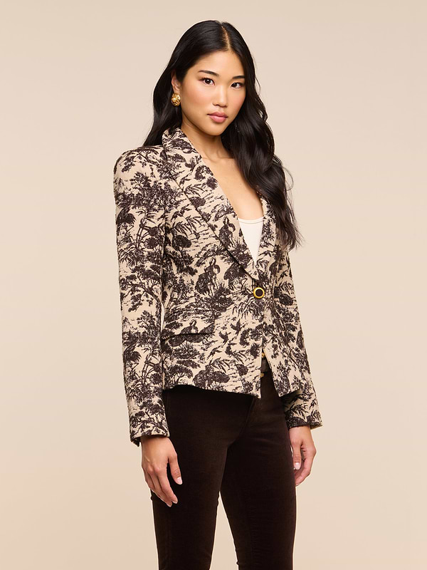 A woman with long black hair wears the Kaisley Blazer, which features an antique-inspired beige and black print, styled over a white top and dark pants as she stands against a plain light background.