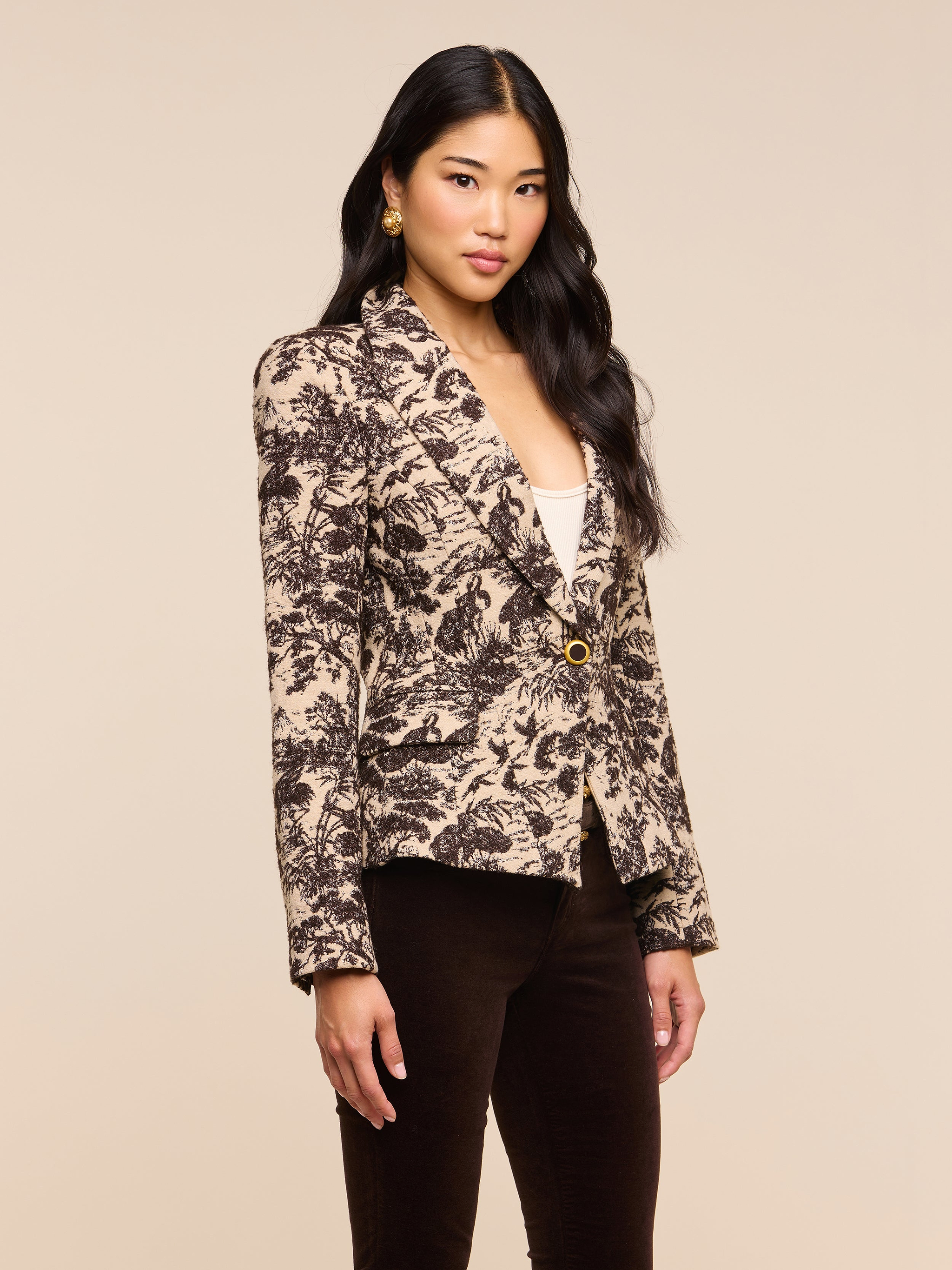 A woman with long black hair wears the Kaisley Blazer, which features an antique-inspired beige and black print, styled over a white top and dark pants as she stands against a plain light background.