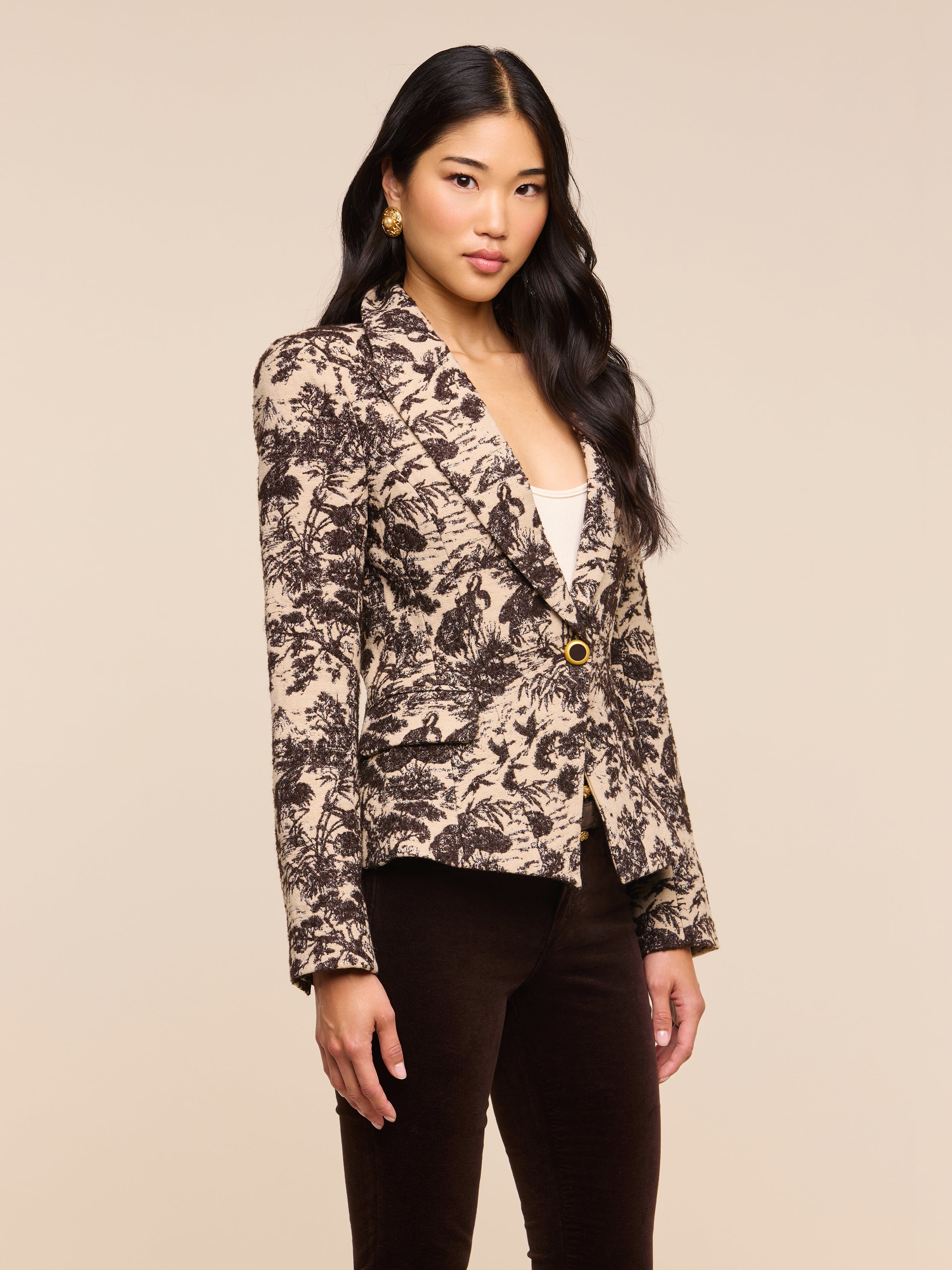 A woman with long black hair wears the Kaisley Blazer, which features an antique-inspired beige and black print, styled over a white top and dark pants as she stands against a plain light background.