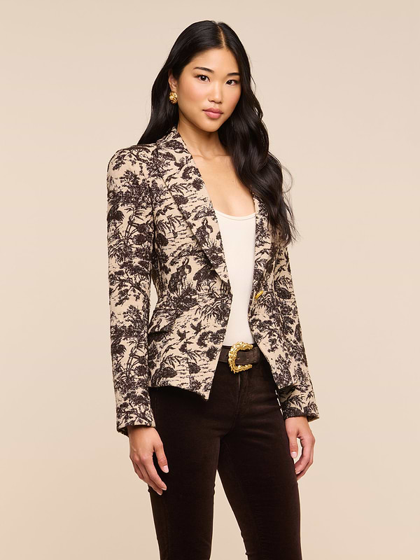 A woman with long dark hair stands confidently against a plain light background, wearing the Kaisley Blazer—an eye-catching piece featuring an antique-inspired print—styled over a light top and dark pants as she looks at the camera.