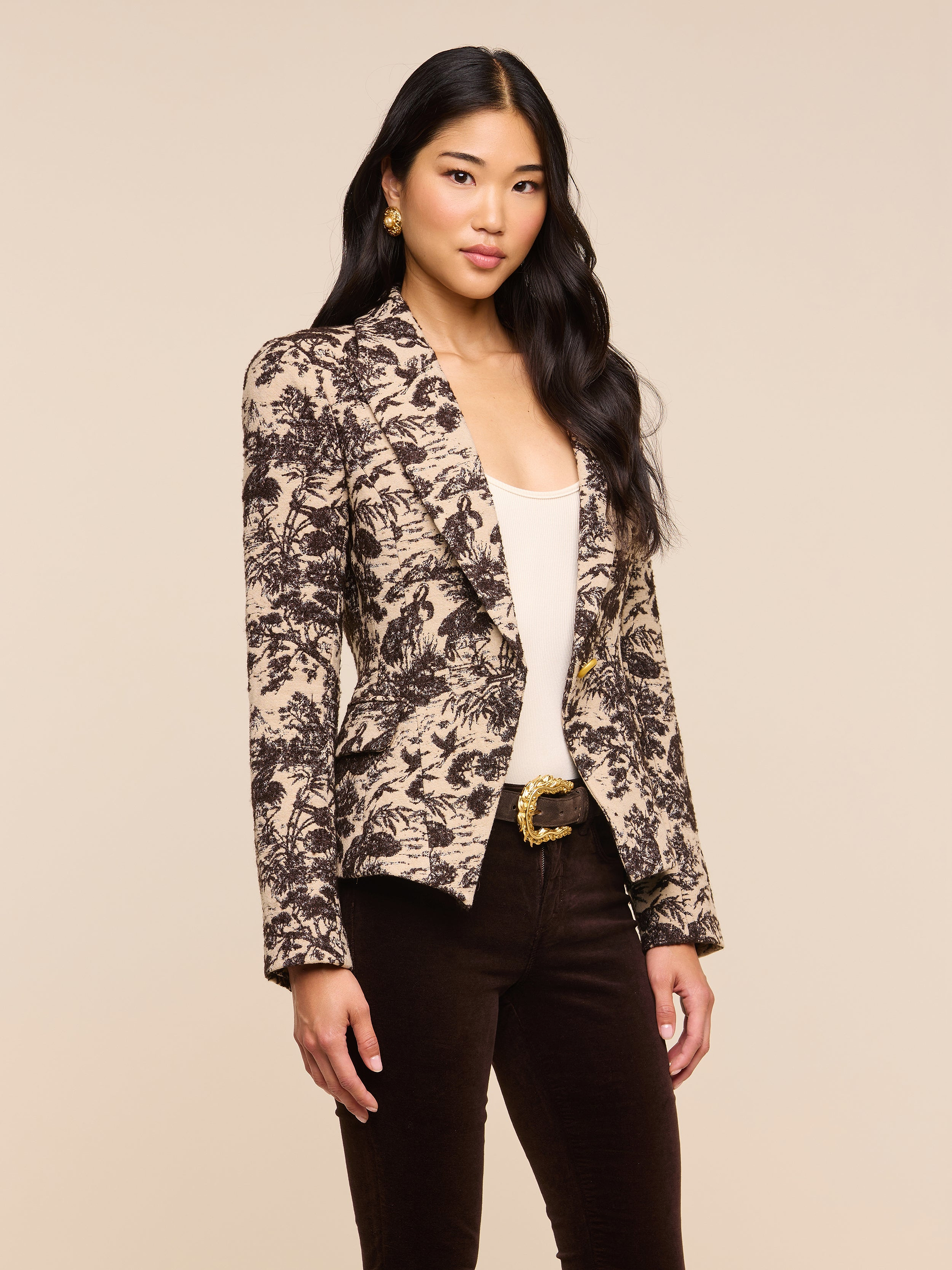 A woman with long dark hair stands confidently against a plain light background, wearing the Kaisley Blazer—an eye-catching piece featuring an antique-inspired print—styled over a light top and dark pants as she looks at the camera.