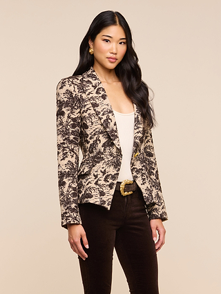 A woman with long dark hair stands against a plain beige background, wearing the Kaisley Blazer—featuring an antique-inspired beige and black print—layered over a white top and paired with dark pants.