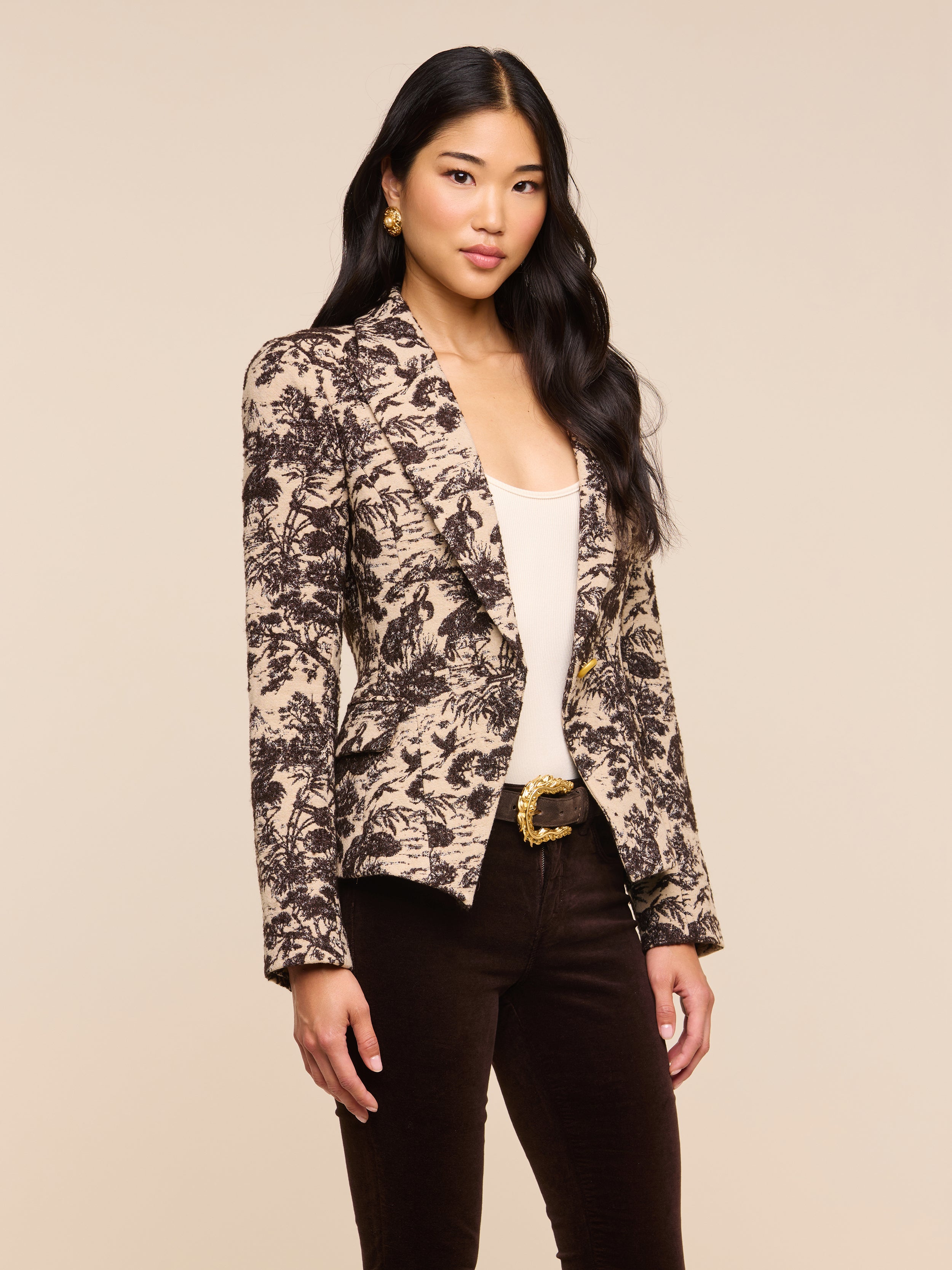 A woman with long dark hair stands confidently against a plain light background, wearing the Kaisley Blazer—an eye-catching piece featuring an antique-inspired print—styled over a light top and dark pants as she looks at the camera.