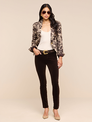 A woman wearing sunglasses, the Kaisley Blazer featuring an antique-inspired print, a white top, dark skinny pants, and beige heels stands against a plain beige background with one hand in her pocket.