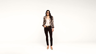 A woman wearing sunglasses, a patterned jacket, white top, black pants, and nude heels stands confidently against a plain white background.