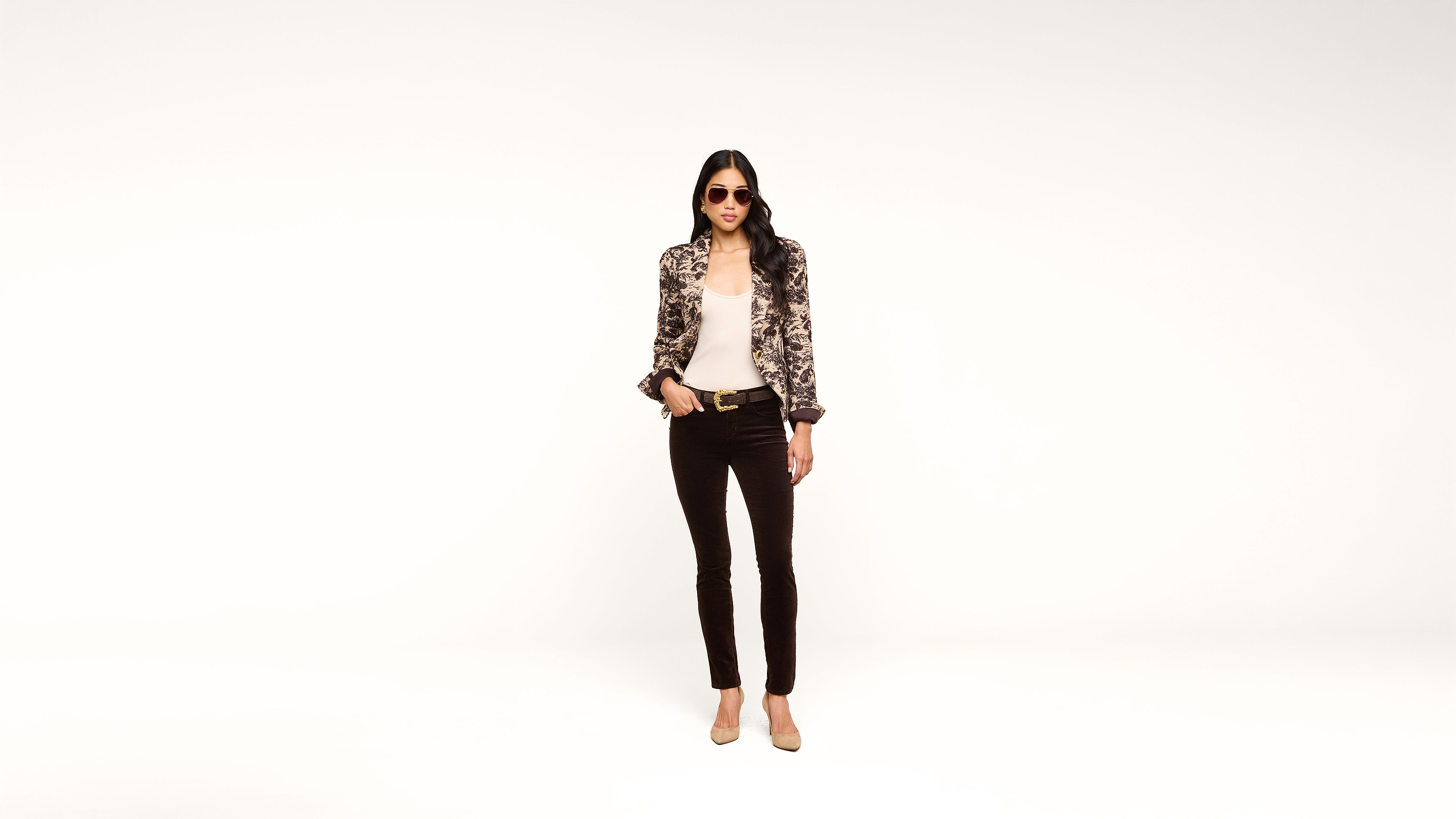 A woman wearing sunglasses, a patterned jacket, white top, black pants, and nude heels stands confidently against a plain white background.