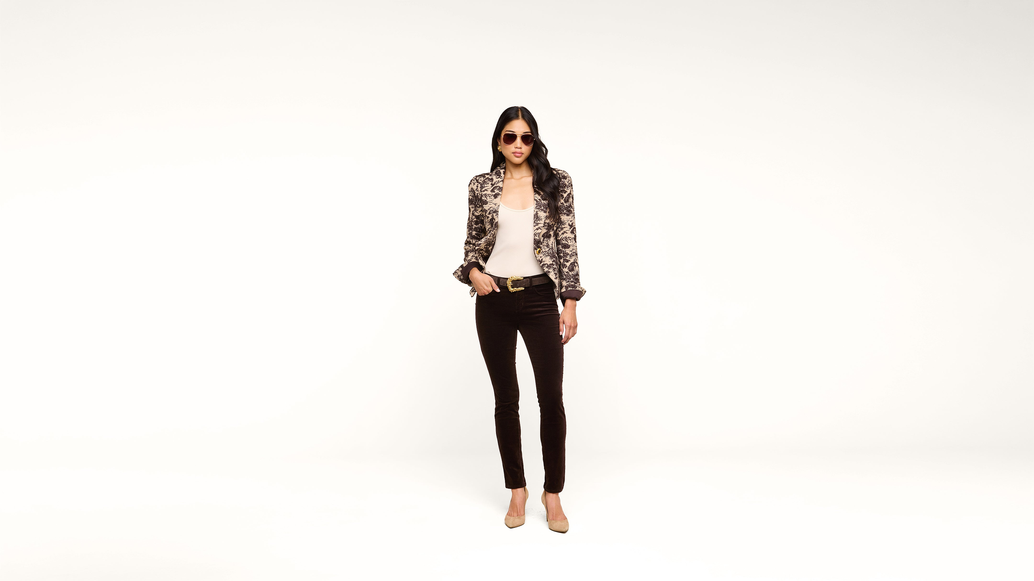 A woman wearing sunglasses, a patterned jacket, white top, black pants, and nude heels stands confidently against a plain white background.