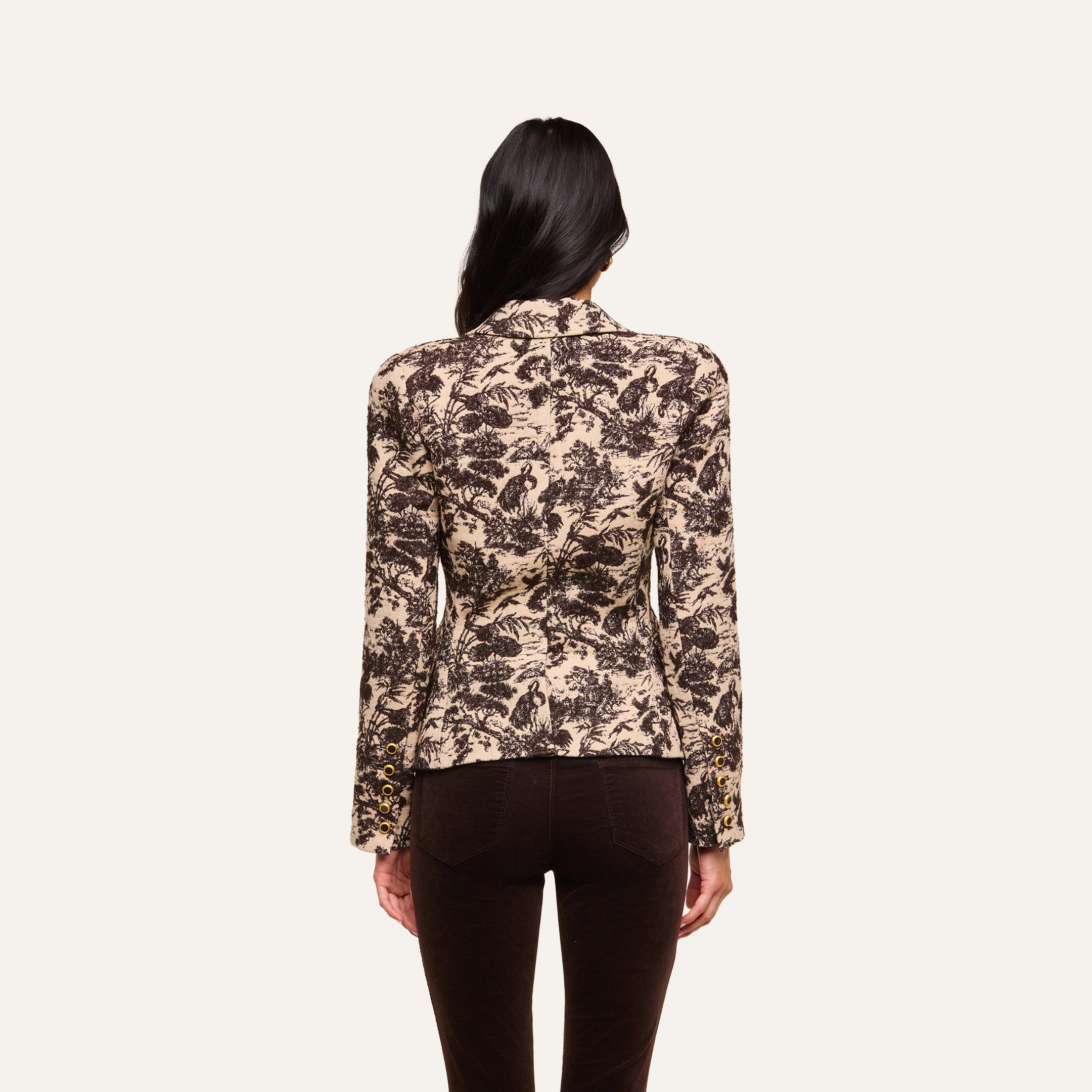 A woman with long dark hair, seen from behind, wears the Kaisley Blazer in beige, featuring an antique-inspired botanical print, paired with dark pants against a plain light background.