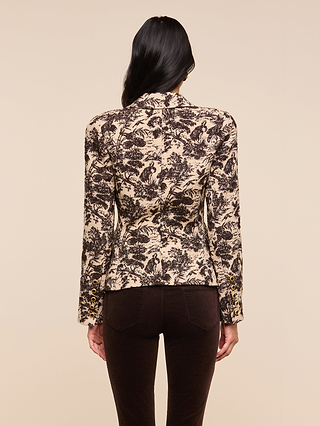 A person with long dark hair is seen from behind, modeling the Kaisley Blazer—which features an antique-inspired beige and black print and gold sleeve buttons—paired with dark brown pants against a plain light background.
